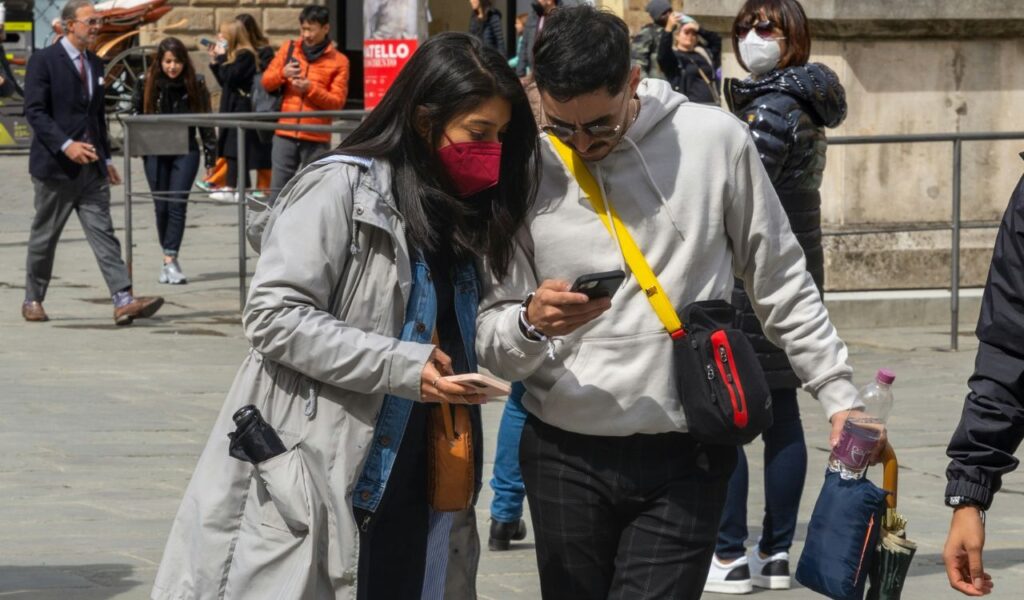 Two tourists wearing casual clothes and masks look at a smartphone together while walking in a busy European square.