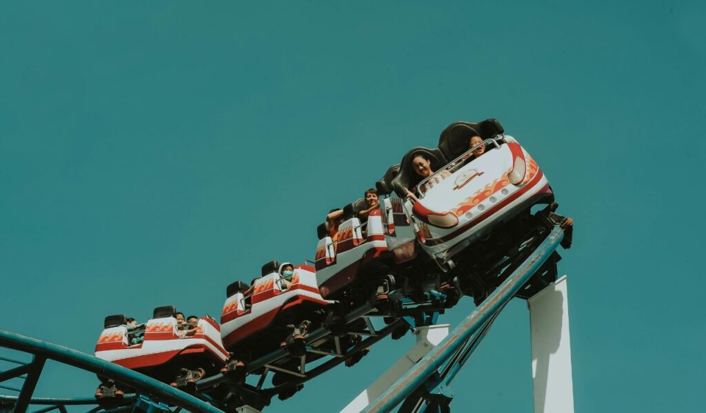 **Alt text:** People riding a colorful roller coaster with flame decals as it twists on a steep track against a clear blue sky.