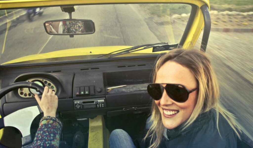 Smiling woman in sunglasses enjoys a ride in a yellow convertible with the road blurring behind, capturing joyful travel.