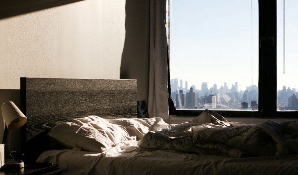 Cozy unmade bed in a modern room with sunlight streaming in, next to a large window offering a clear view of the New York City skyline in the background.
