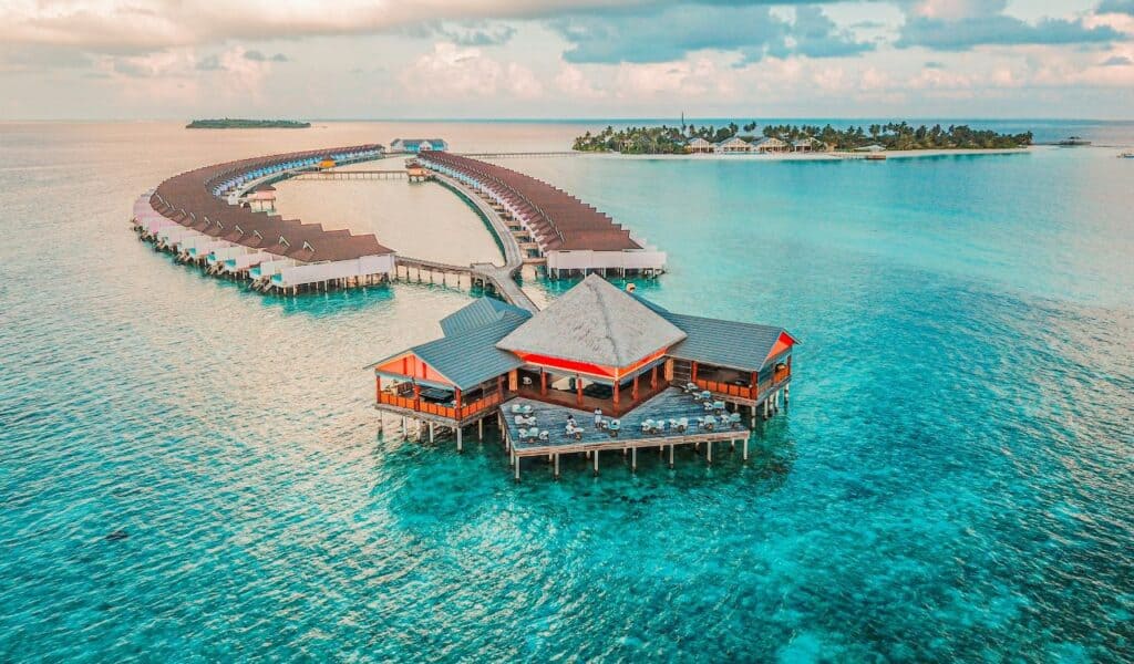 Aerial view of overwater villas and a seaside restaurant at a luxury resort in the Maldives, surrounded by clear turquoise ocean and tropical islands in the background.