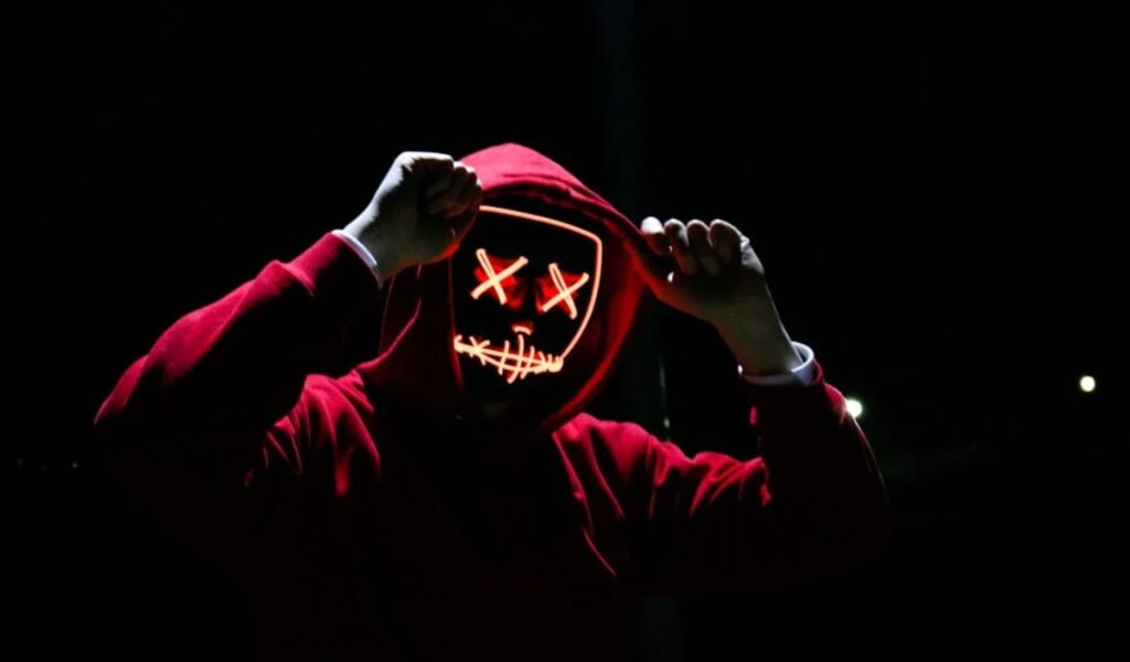 A person in a red hoodie wearing a glowing neon mask with stitched lips and crossed-out eyes, standing in the dark, suggesting a mysterious or potentially deceptive street performer.