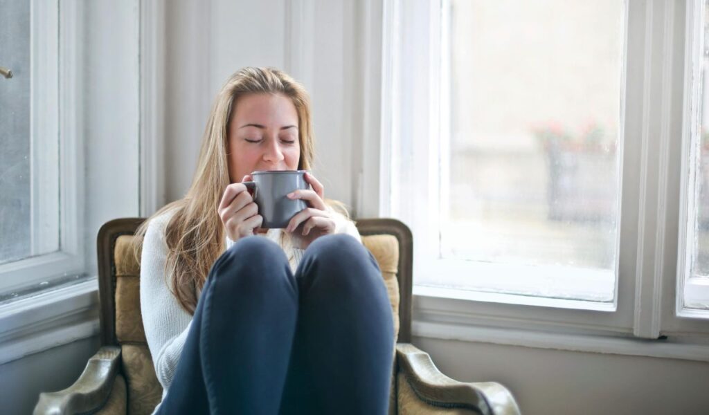 A woman sits comfortably in a cozy chair by a window, eyes closed and smiling softly as she holds a warm mug in both hands.