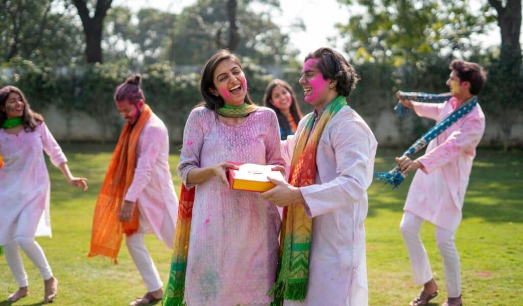 Group of people celebrating Holi outdoors, wearing colorful traditional clothes and smeared with bright powder, smiling and exchanging a gift.