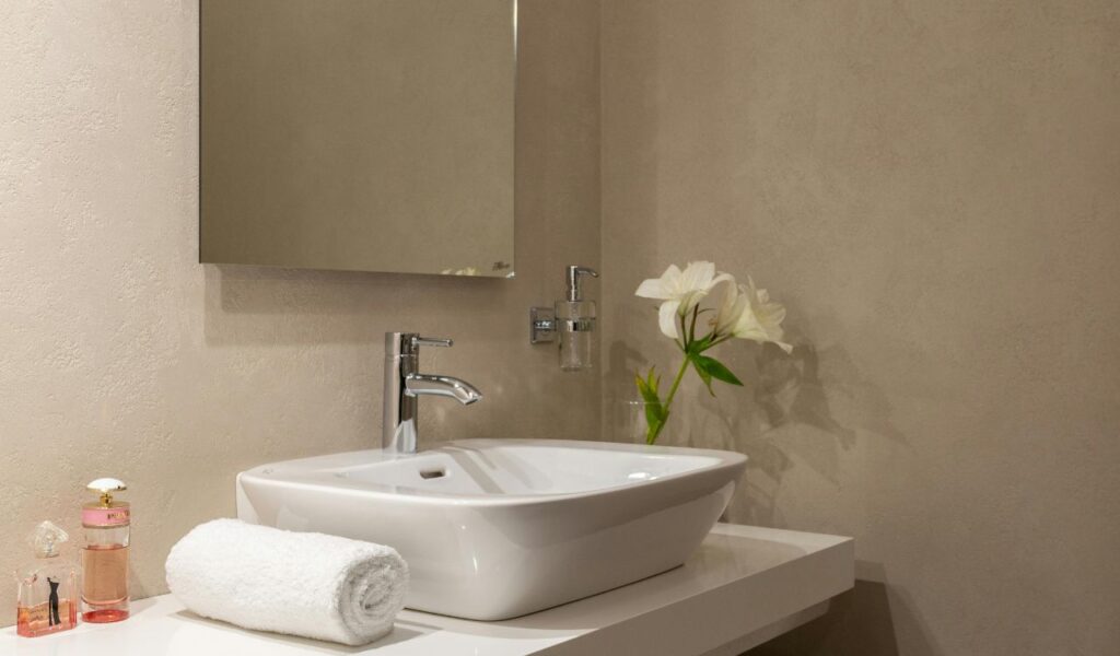 Modern hotel bathroom sink with a rolled white towel, perfume bottles, and a vase of white lilies.