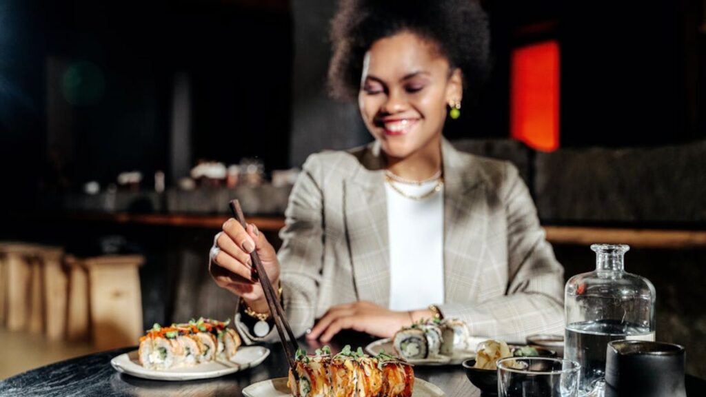 A smiling woman uses chopsticks to pick up sushi from a plate at a restaurant table, with various sushi dishes and drinks in front of her, representing dining etiquette and enjoyment of Japanese cuisine.