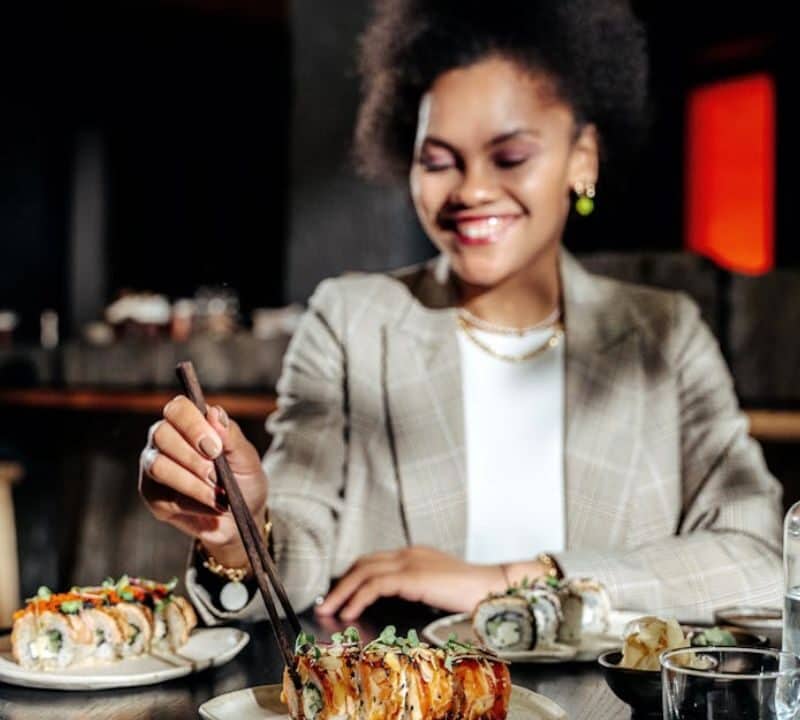 A smiling woman uses chopsticks to pick up sushi from a plate at a restaurant table, with various sushi dishes and drinks in front of her, representing dining etiquette and enjoyment of Japanese cuisine.