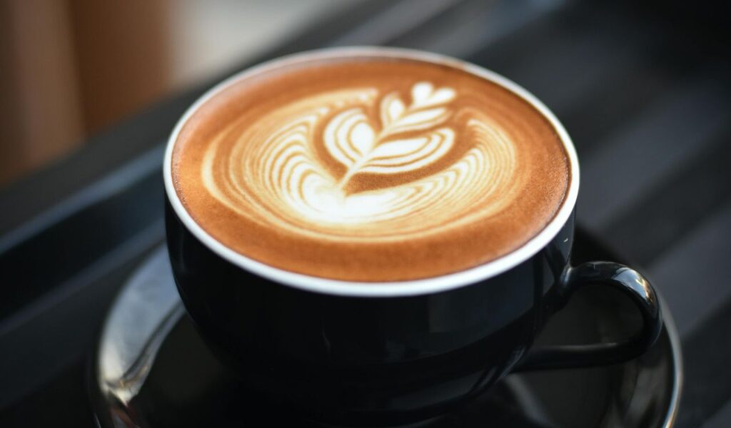 A close-up of a black cup of coffee with intricate latte art on top, placed on a matching saucer against a blurred background.
