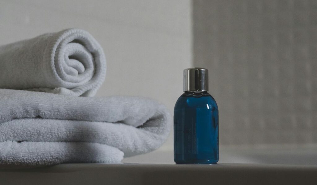 Rolled white towels stacked neatly beside a small blue bottle of toiletry on a bathroom surface with soft lighting.