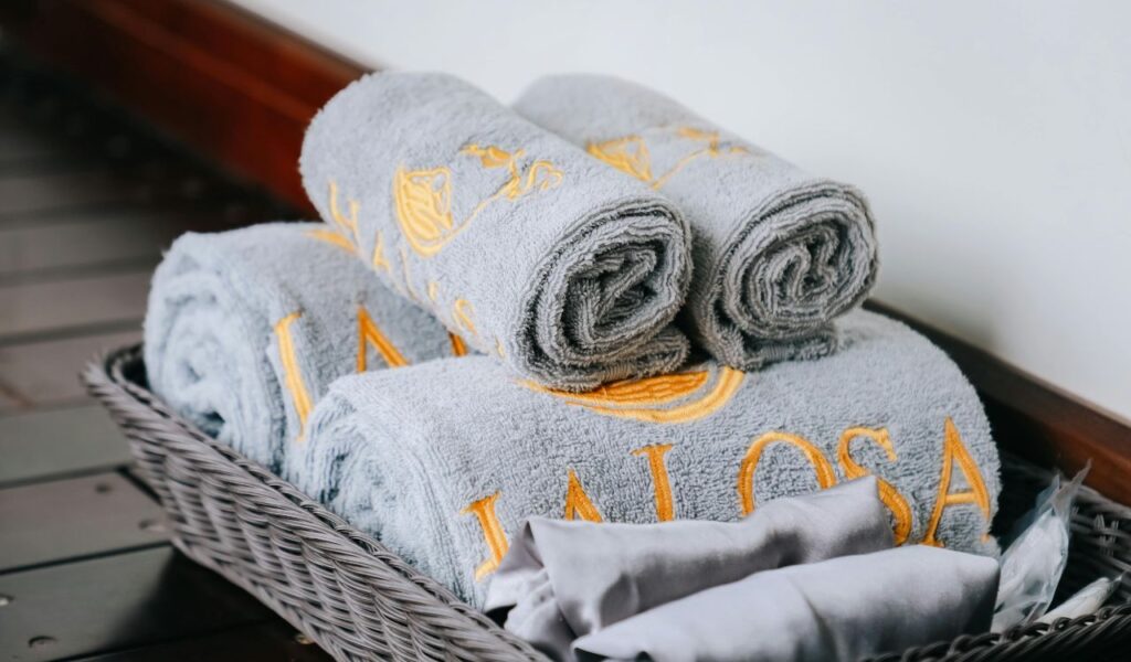 Rolled gray hotel towels with yellow embroidery neatly arranged in a wicker basket on a wooden surface.