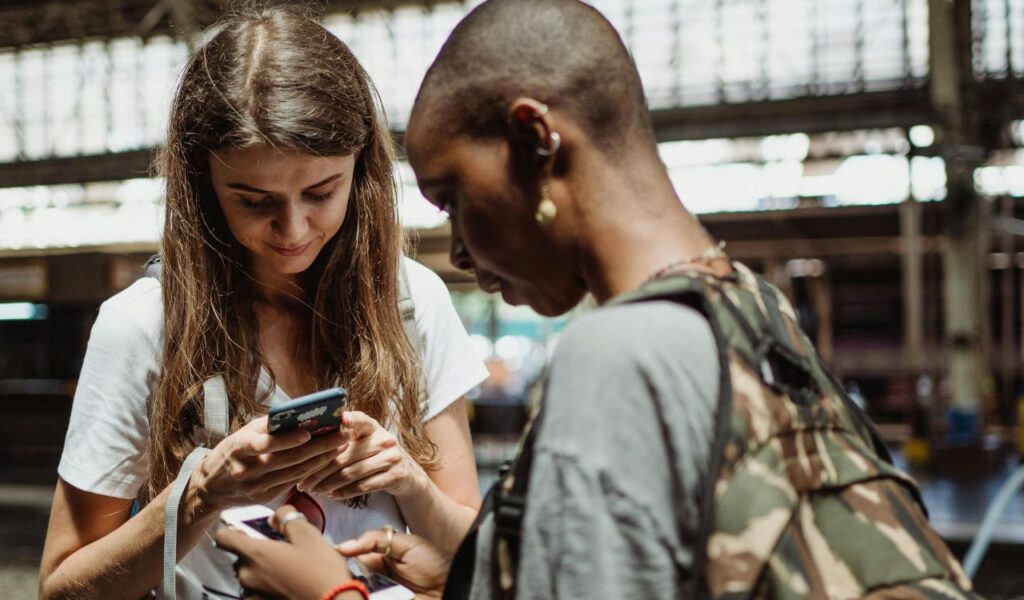 Two travelers stand inside a train station looking at their phones, one in a camo backpack and the other with a white t-shirt and long brown hair.