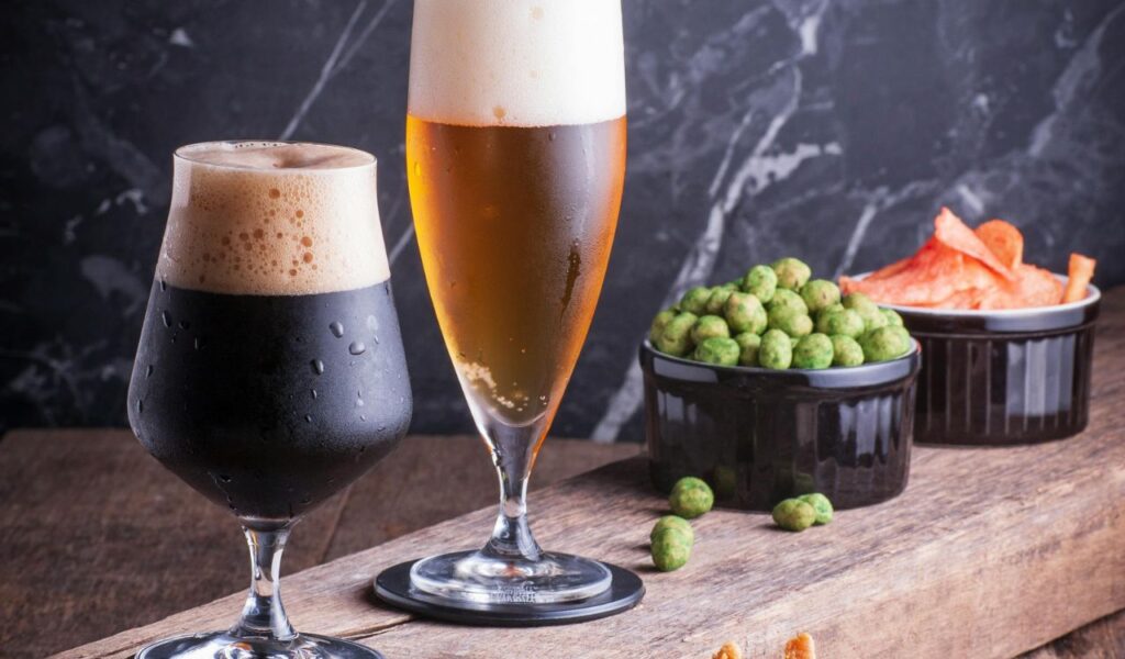 Two glasses of beer, one dark stout and one golden lager, served with bowls of green peas and potato chips on a rustic wooden table.