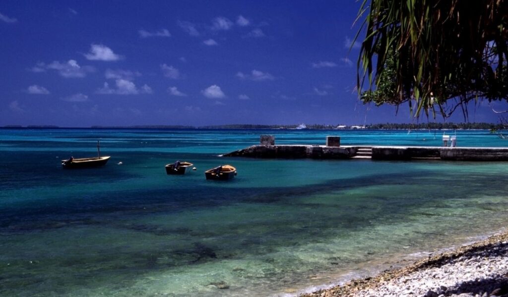 Crystal-clear lagoon in Tuvalu with small boats, stone pier, and vibrant blue water under a deep blue tropical sky.