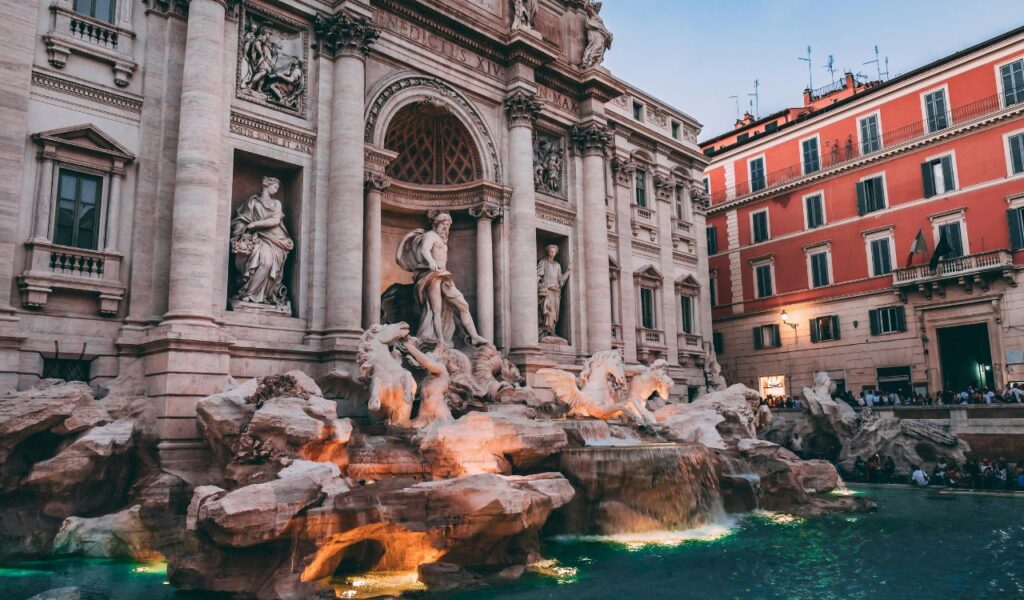 Trevi Fountain in Rome illuminated at dusk, featuring Neptune statue, rearing horses, and cascading turquoise water.
