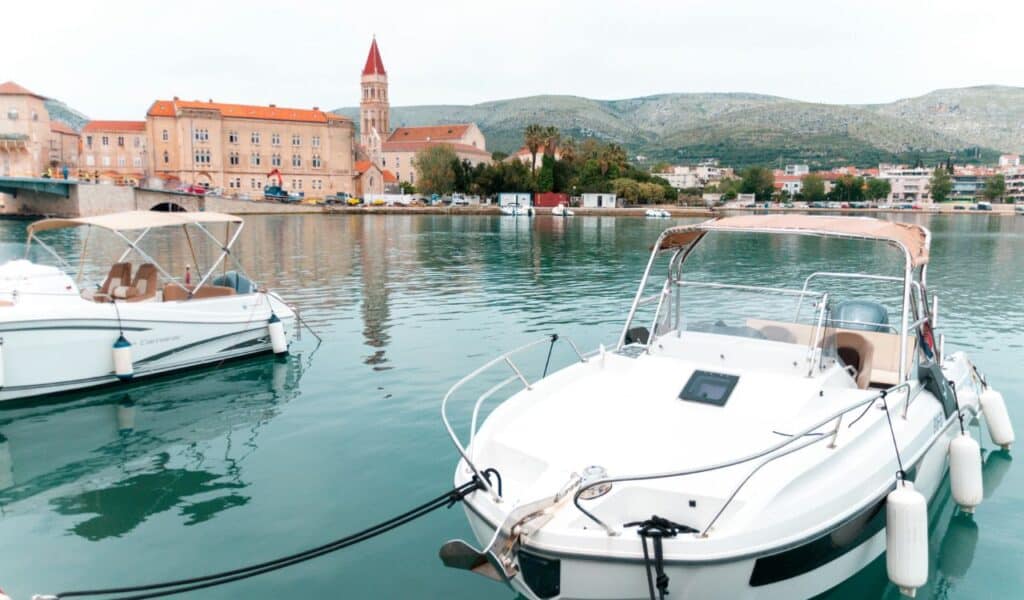 Alt text:
Small motorboats docked in the calm turquoise waters of Trogir’s harbor, with a red-roofed historic building and church tower in the background, set against green hills.