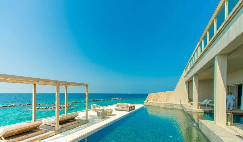 A luxury oceanfront resort features an infinity pool, shaded lounge chairs, and a clear view of turquoise waters under a bright blue sky.