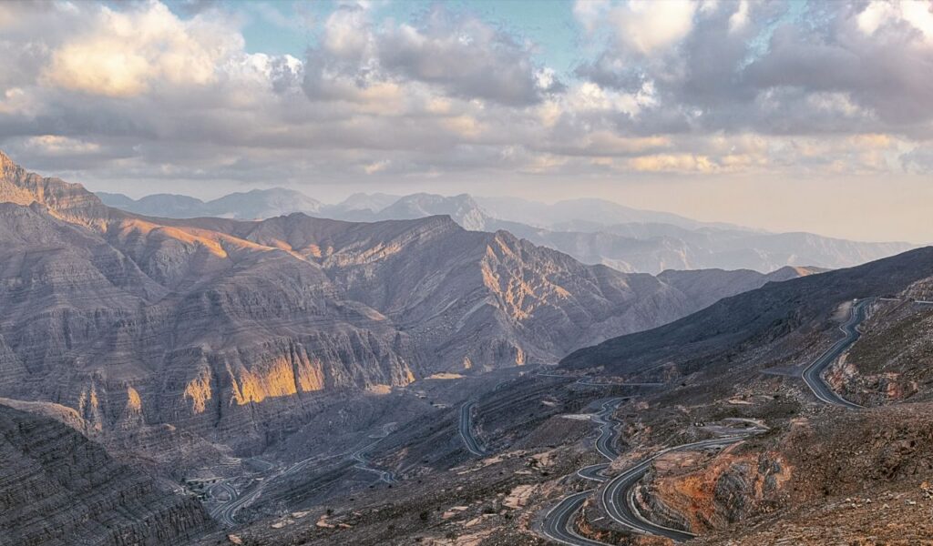 Winding mountain roads cutting through the rugged Hajar Mountains in the United Arab Emirates under a partly cloudy sky.