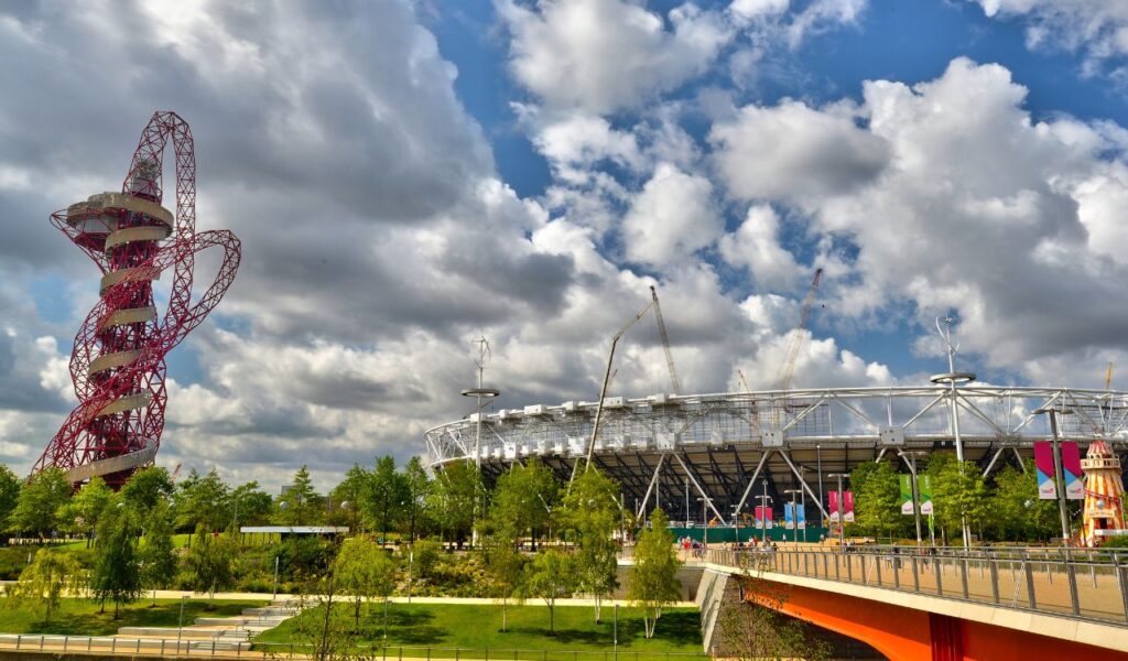 The ArcelorMittal Orbit, a twisting red steel sculpture with observation decks, stands beside the London Olympic Stadium surrounded by greenery and flags.