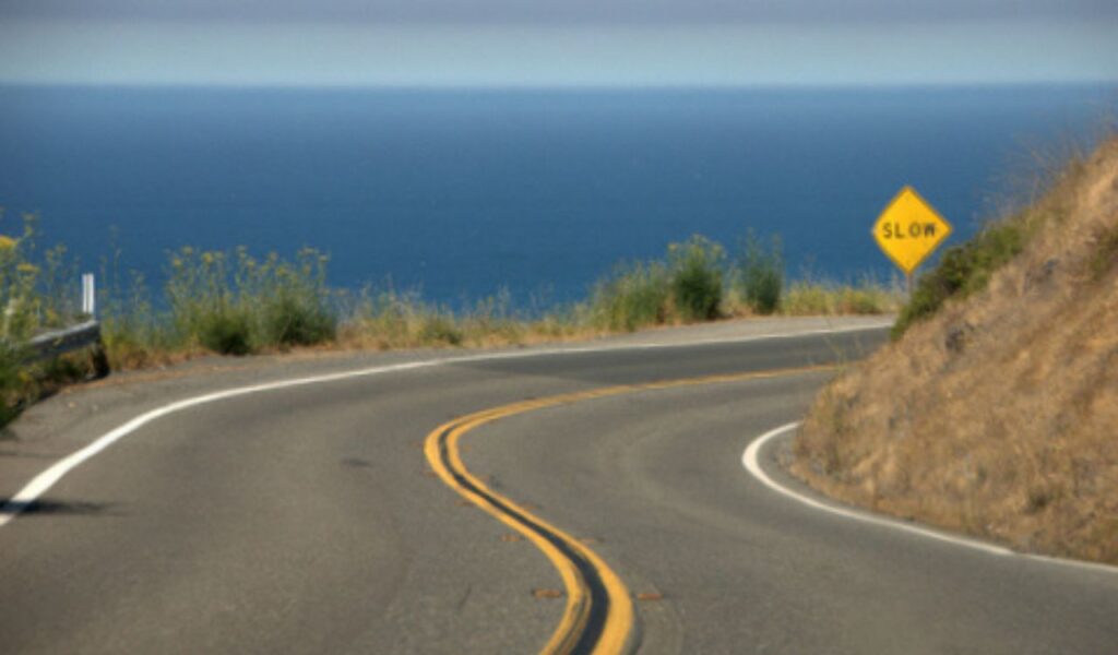 Winding stretch of California’s Pacific Coast Highway with ocean views and a “Slow” caution sign on a sunny day.
