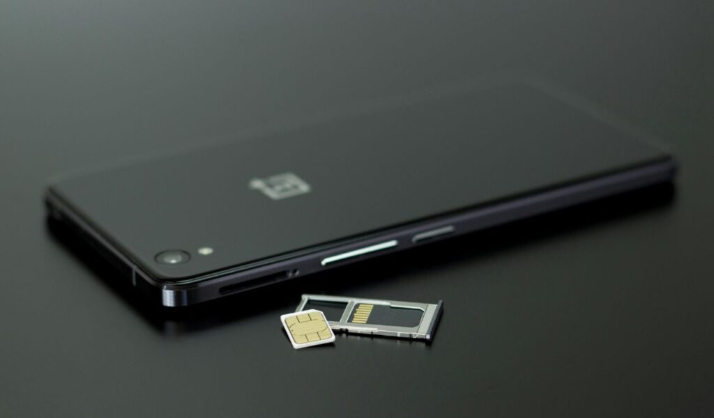 A black smartphone with a SIM card tray and a local SIM card placed beside it on a dark surface.
