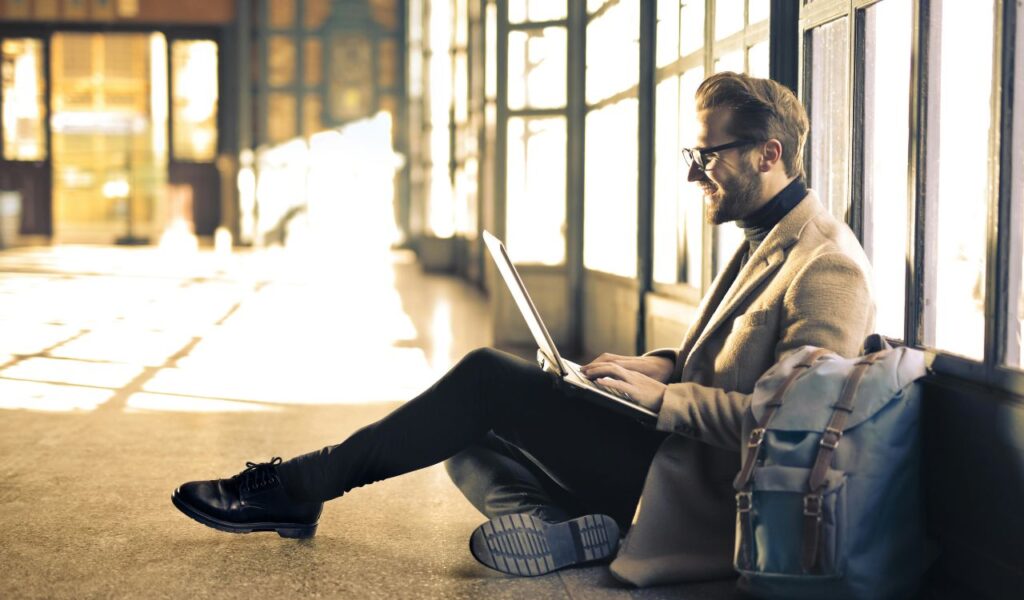 Smiling man sitting on the floor in a sunlit terminal using a laptop, with a travel backpack beside him.