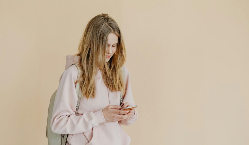 Young woman wearing a pink hoodie and backpack looking down at her smartphone while standing against a plain beige background.