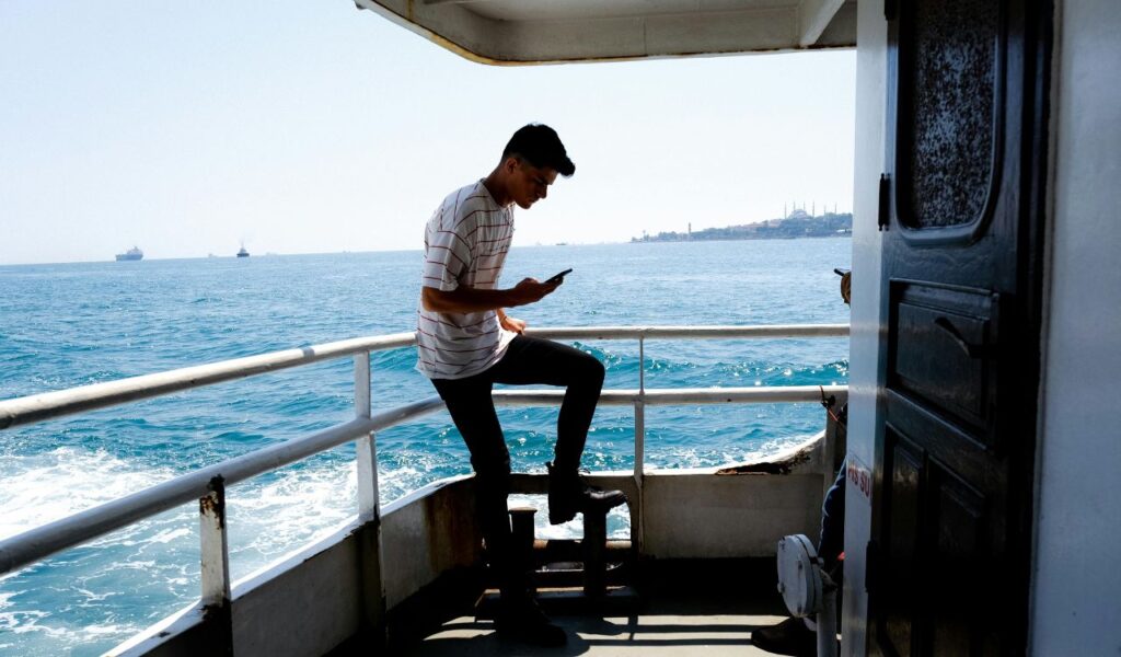 Man standing on the deck of a boat, looking at his phone with the ocean and distant ships in the background on a sunny day.