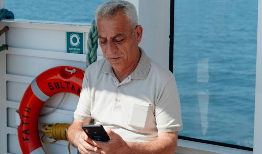 An elderly man using a smartphone while sitting on a boat with a lifebuoy in the background.