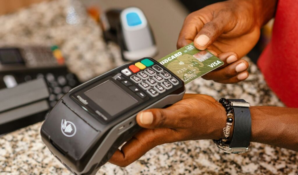 A person holds a green Visa credit card near a contactless payment terminal on a granite counter, preparing to make a purchase.