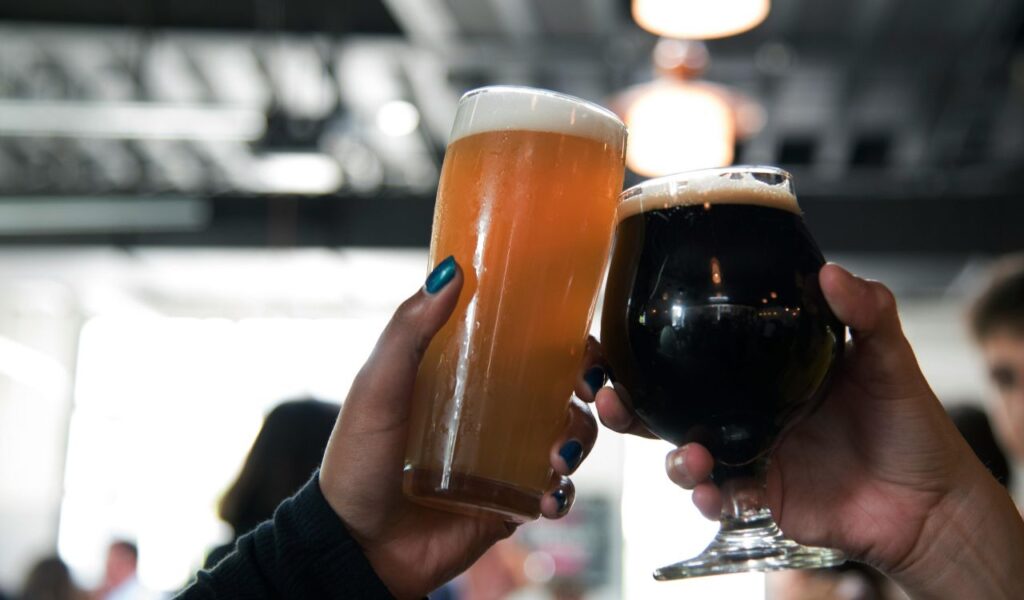 Two people clinking glasses of beer, one with a light lager and the other with a dark stout, in a lively pub setting.