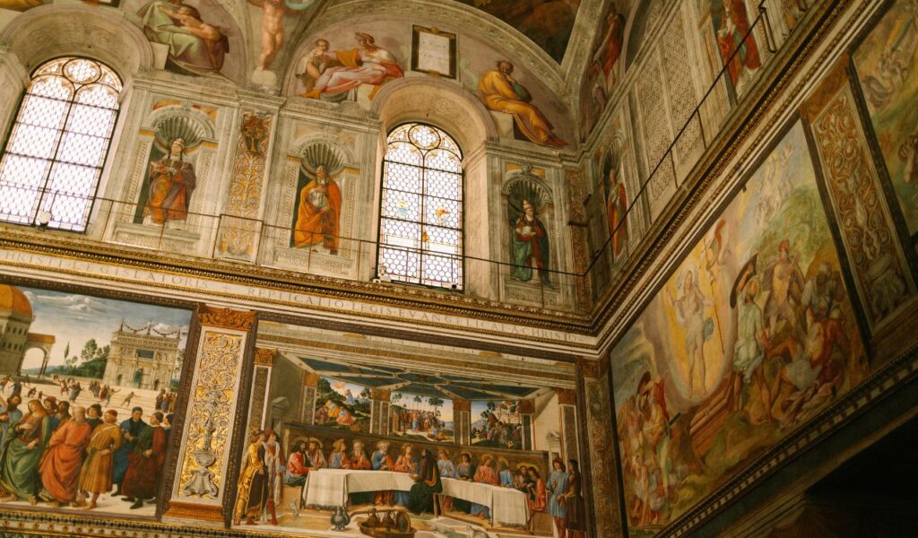 Interior view of the Vatican Museums showing ornate frescoes, religious scenes, arched windows, and Renaissance art.