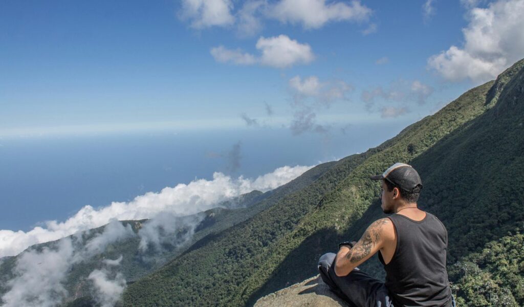 A man sitting on a rocky ledge overlooking lush green mountains and a vast blue sky in Venezuela, with clouds drifting below.