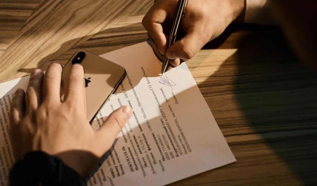 A close-up of hands signing a document on a wooden desk, with a smartphone placed nearby, representing the process of reviewing and verifying official paperwork or permits.