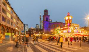 Alt text: Vibrant Christmas market in Frankfurt with crowds walking past brightly lit stalls and a large decorated clock tower, set against a backdrop of historic and modern buildings at dusk.