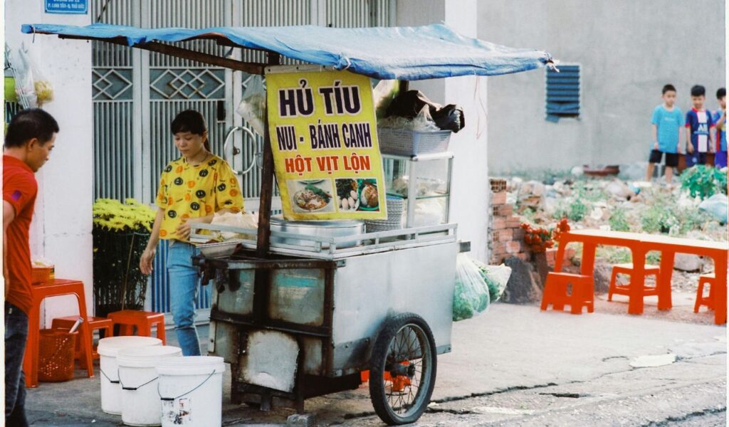 Vietnamese street food vendor selling Hủ Tiếu noodles and duck eggs from a traditional food cart in a local neighborhood.