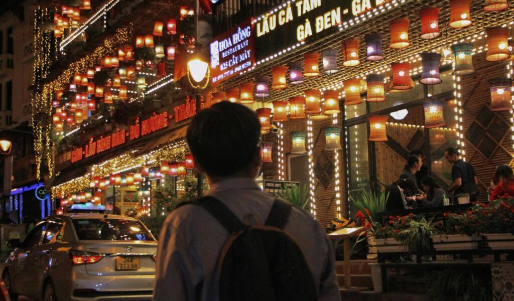 Backpacker stands in front of a brightly lit Vietnamese restaurant at night, surrounded by colorful lanterns and busy street life.
