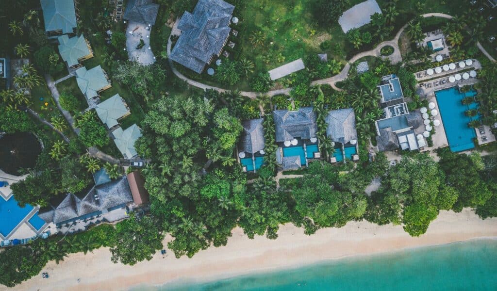 Aerial view of beachfront villas nestled among lush greenery, with private plunge pools, winding paths, and a turquoise shoreline, showcasing a luxury tropical resort setting.