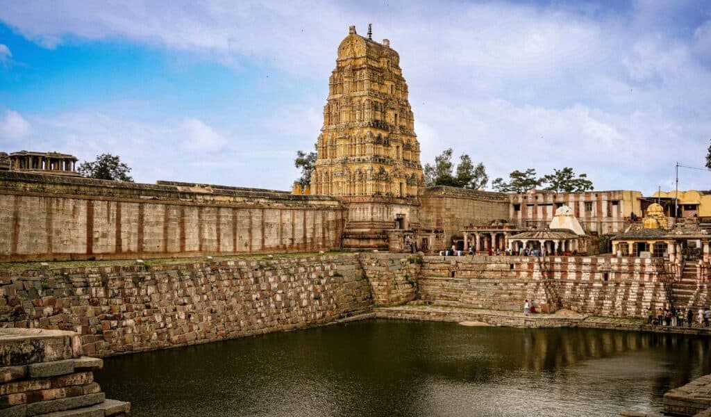Ancient temple complex with a tall, intricately carved gopuram (gateway tower) reflecting in a large stepped water tank, surrounded by stone walls and smaller shrines, under a partly cloudy sky.