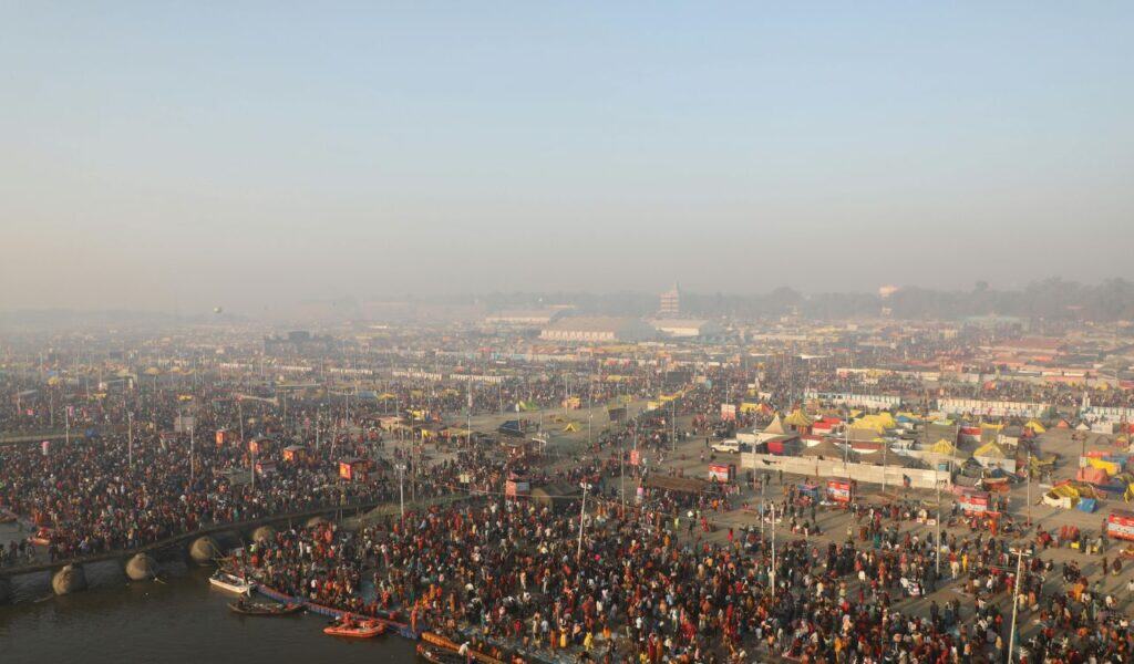Massive crowd gathered at the Sangam in Prayagraj for a religious festival, with colorful tents, boats, and temporary structures spread across the riverbanks under a hazy sky.