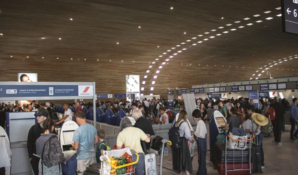Crowded airport check-in area with long lines of travelers, luggage carts, and staff assisting passengers at counters.