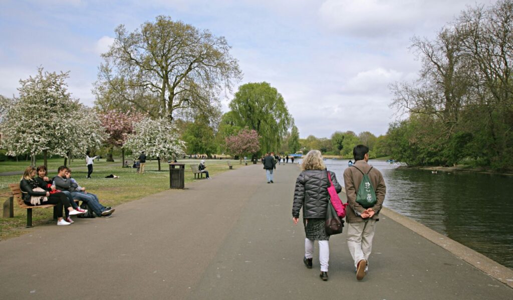 People walk along a tree-lined path beside a calm lake in a city park, with spring blossoms, benches, and others relaxing or strolling nearby.