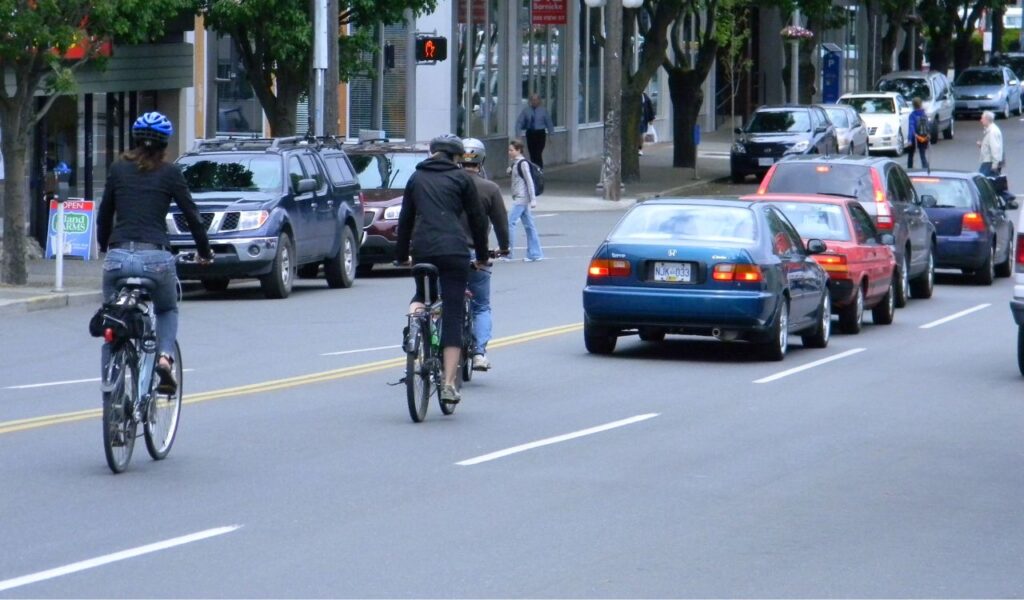 Cyclists riding alongside traffic in a busy urban street while pedestrians walk nearby, highlighting road awareness.