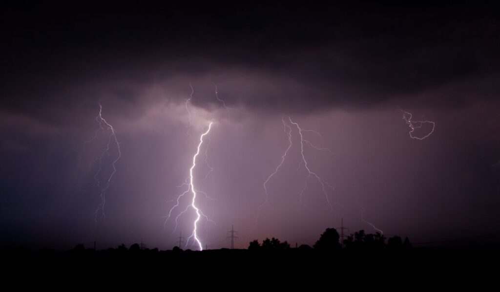 Multiple lightning bolts strike during a dark thunderstorm over a rural landscape, signaling dangerous severe weather.