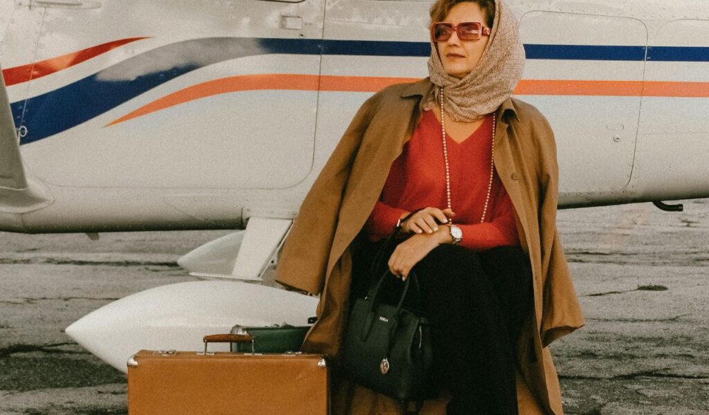 Woman in layered travel outfit with coat and scarf, sitting by airplane with suitcase and handbag, ready to fly.