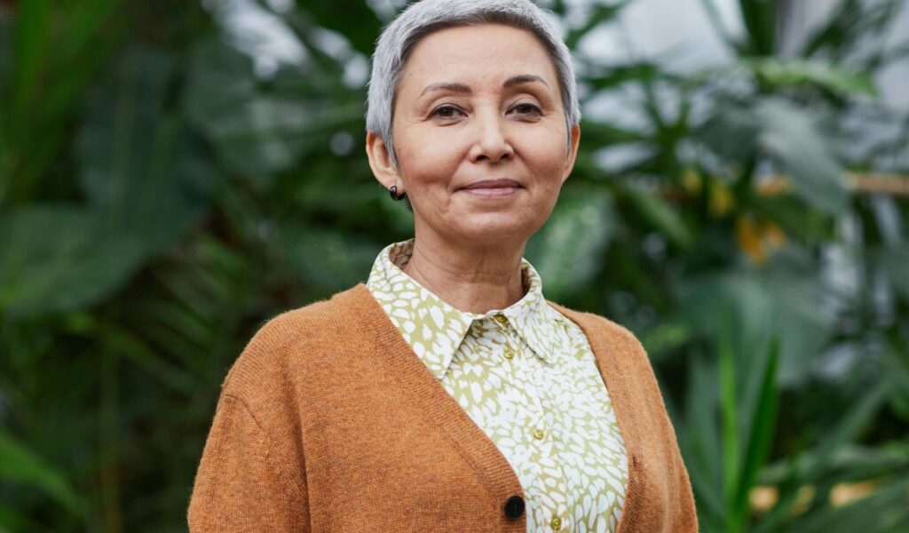 A confident older woman with short gray hair smiles gently, wearing a patterned blouse and a cozy brown cardigan, standing outdoors with lush green foliage in the background.