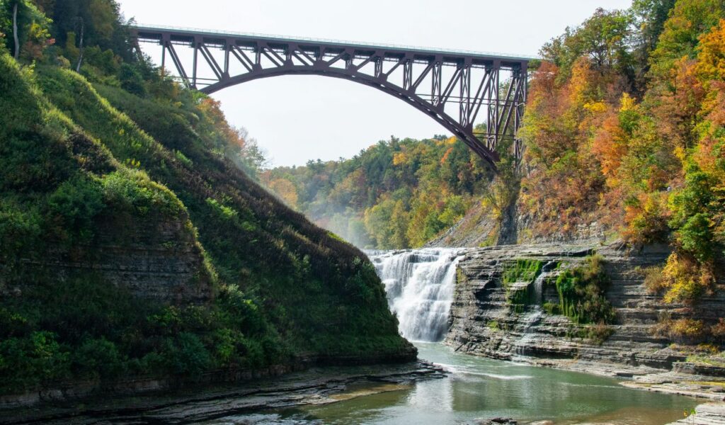 A tall metal bridge spans a deep gorge above a cascading waterfall surrounded by layered rock formations and autumn-colored trees in West Virginia