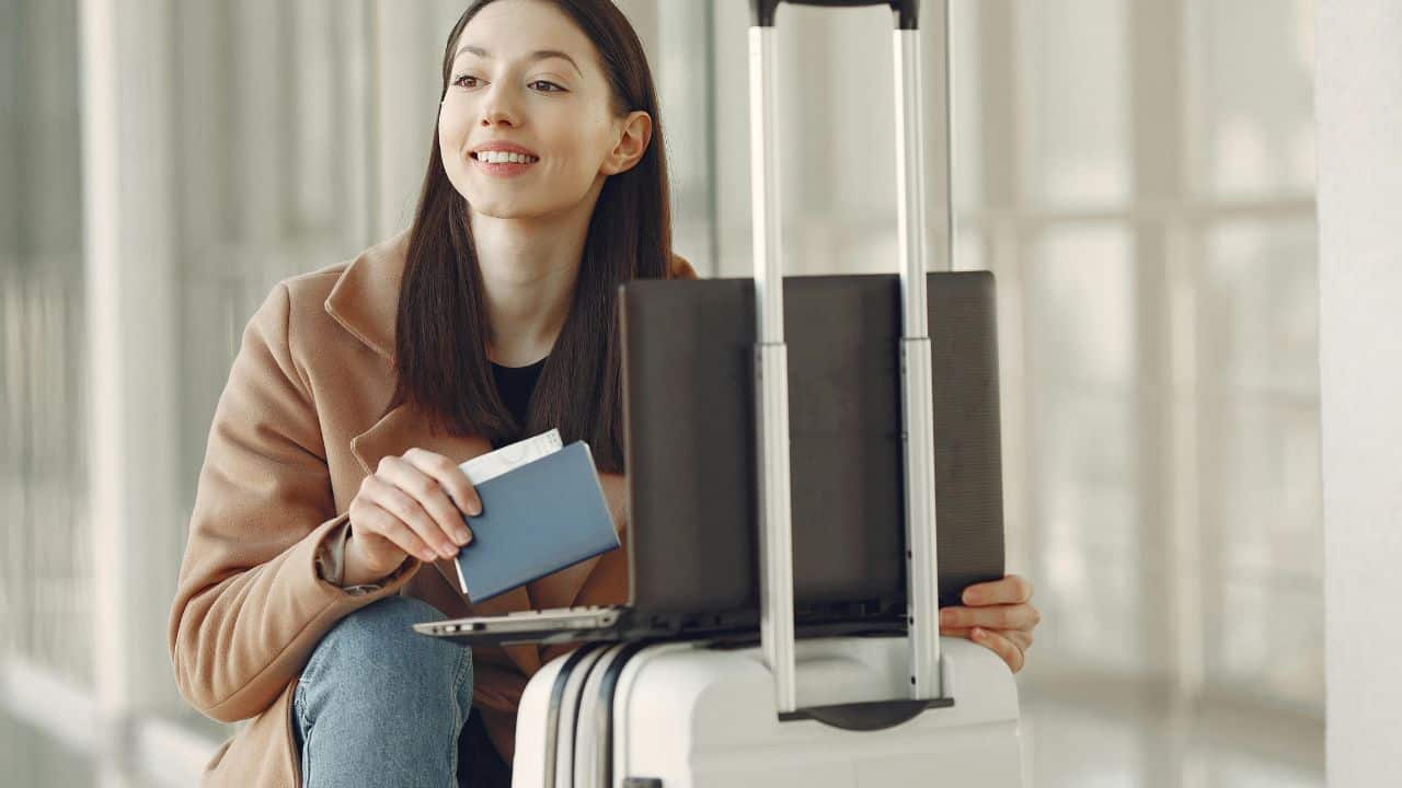**Alt text:** A woman sitting in an airport terminal, smiling while holding a passport and boarding pass. She has a laptop placed on top of her suitcase and appears ready to travel.