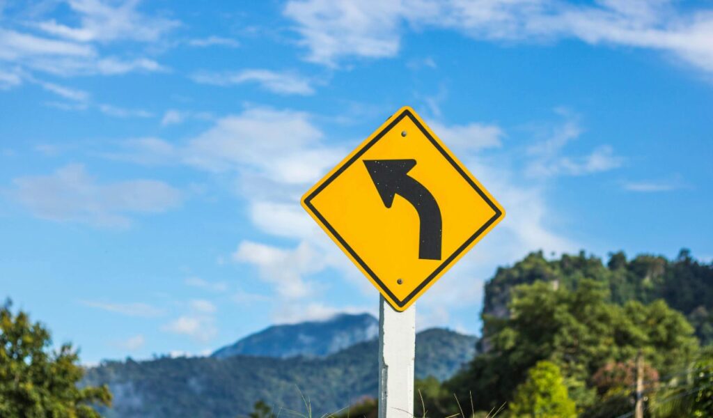 A bright yellow road sign with a black arrow indicates a sharp left curve ahead, set against a backdrop of blue sky and forested mountains.