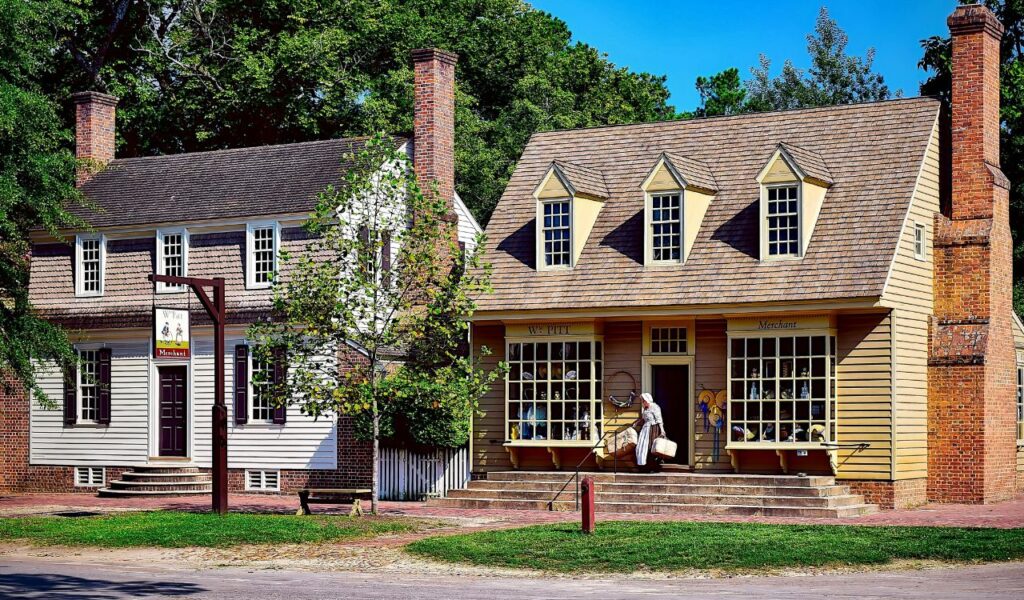 Historic Colonial Williamsburg street with 18th-century buildings, brick chimneys, and a costumed person entering the W. Pitt Merchant shop.