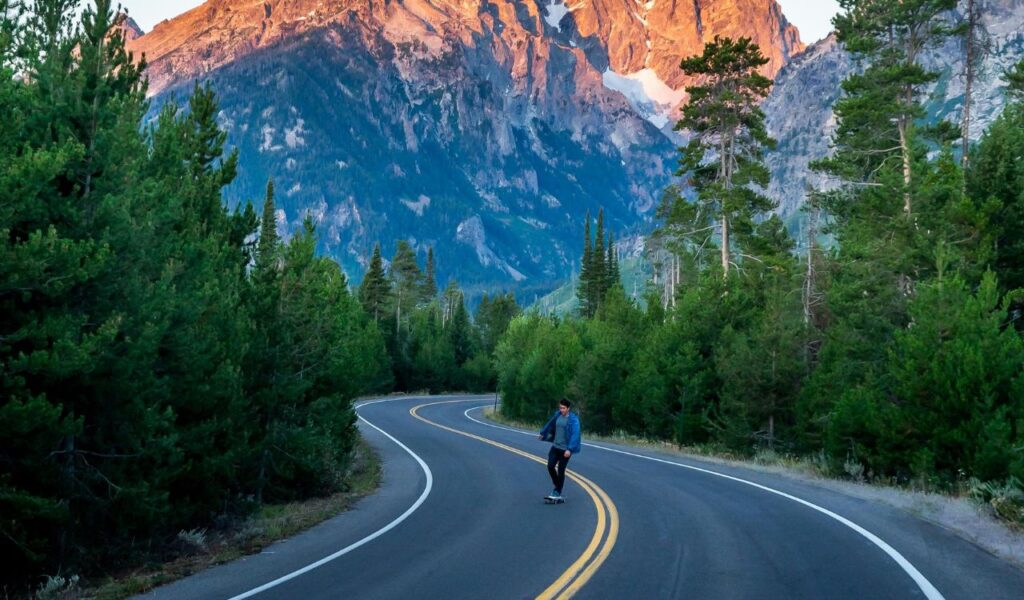 A skateboarder rides along a winding mountain road surrounded by dense pine trees, with the towering, sunlit peaks of the Grand Tetons rising dramatically in the background.